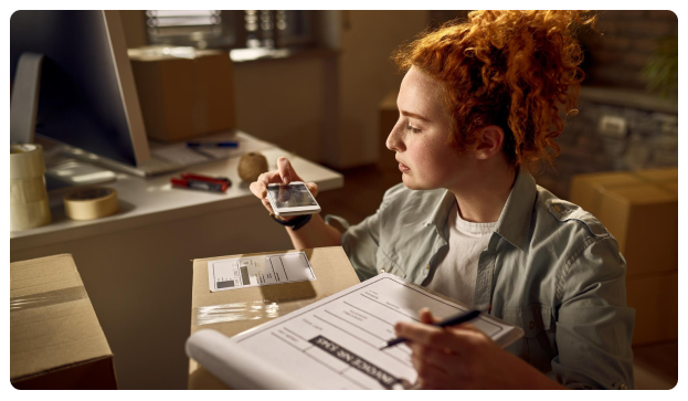 Woman with curly red hair scanning a package with a phone while holding a clipboard
